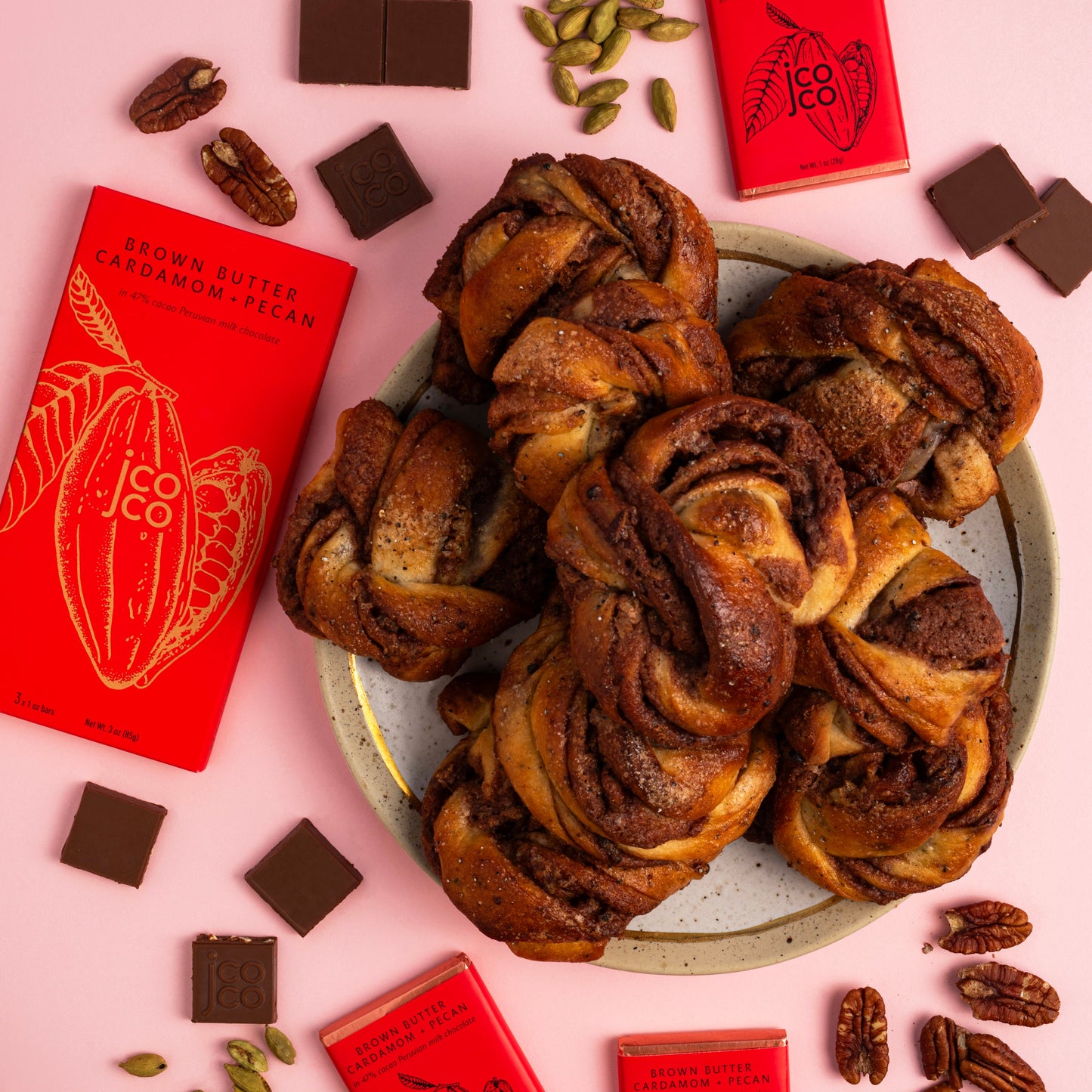 Braided bread with chocolate swirls on a plate, surrounded by red packets and chocolate pieces on a pink background.