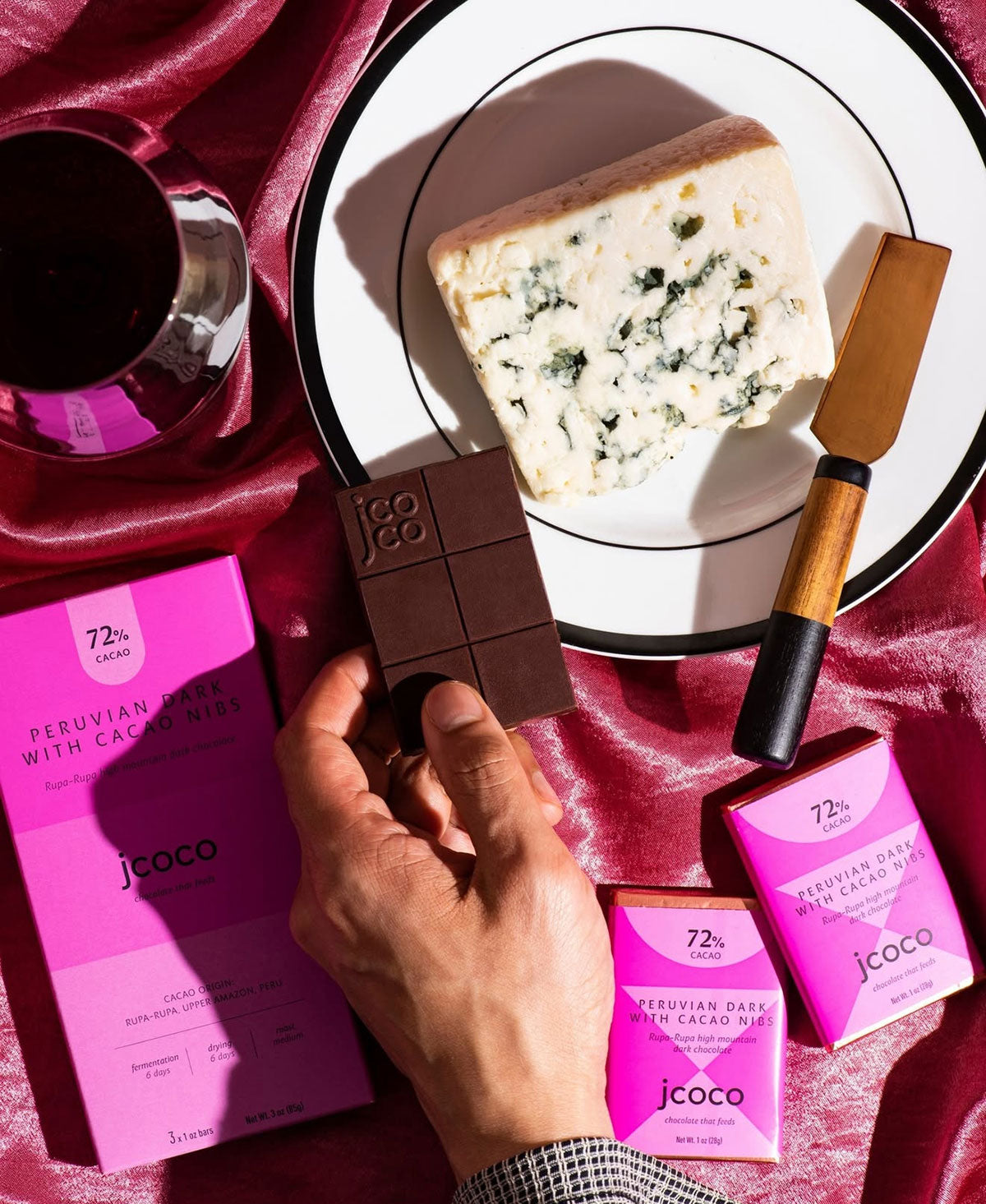 Hand holding a piece of Peruvian chocolate with jcoco packaging, next to a plate of blue cheese and a glass of red wine on a pink surface.